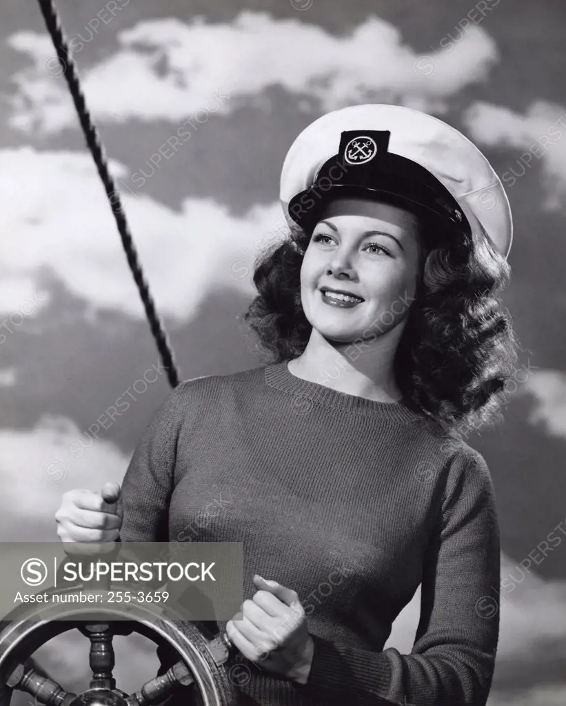 Vintage Photograph. Smiling brunette woman wearing sweater and captain's hat in front of cloud sky background standing with boat helm steering wheel