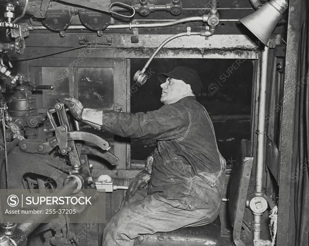 Vintage photograph. Locomotive engineer sitting at controls in his cab and talking to the control tower through two way radio device. Microphone can be seen hanging above his head and speaker above and to the rear of him
