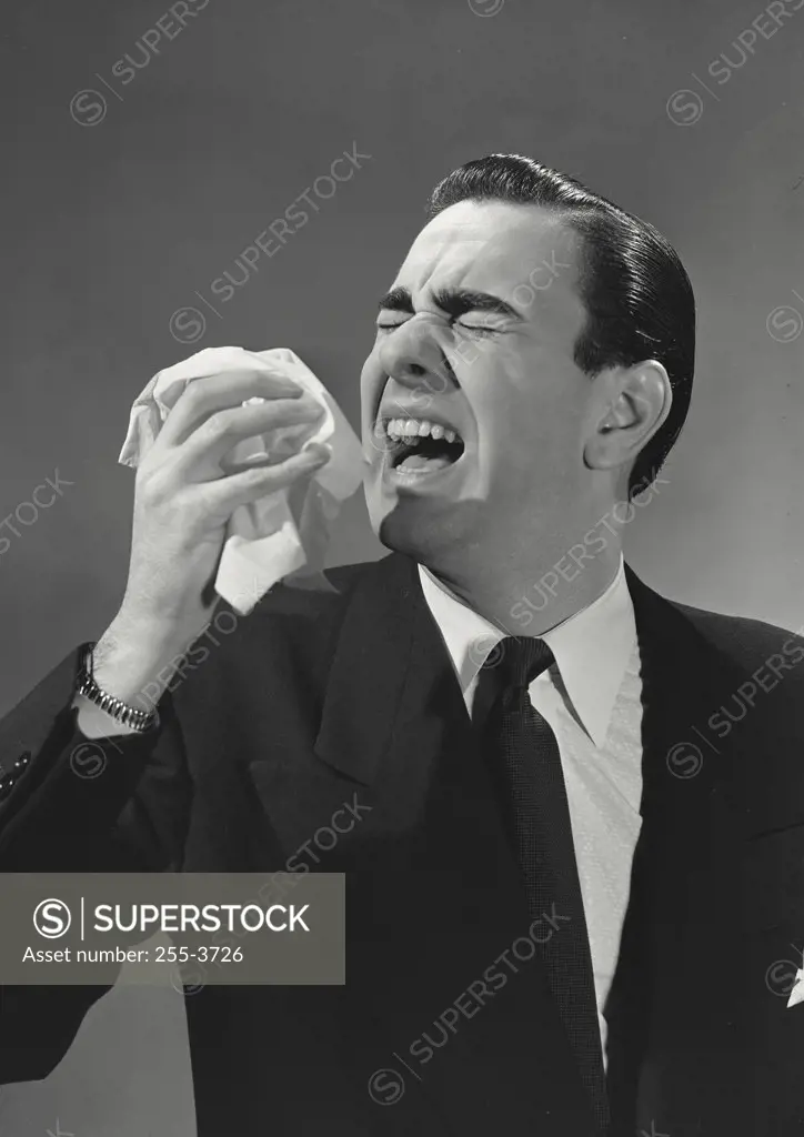 Man with black slicked back hair wearing suit and tie sneezing