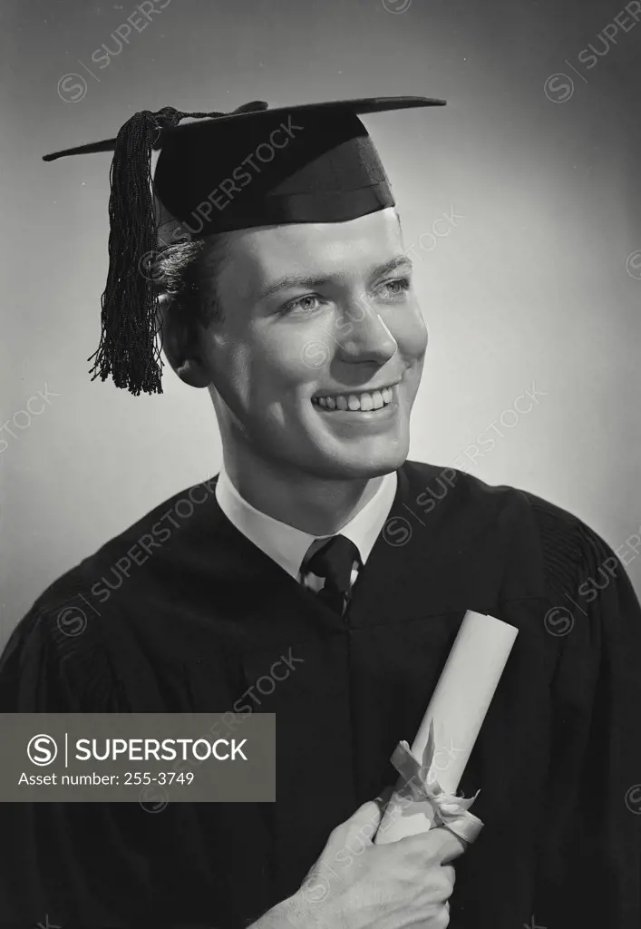 Vintage photograph. Portrait of man in cap and gown holding rolled diploma looking off camera