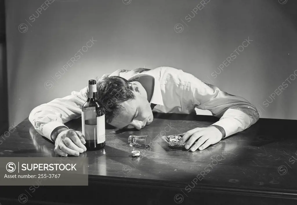Vintage photograph. Drunk man passed out with whiskey bottle and shot glass on table with hand in ashtray