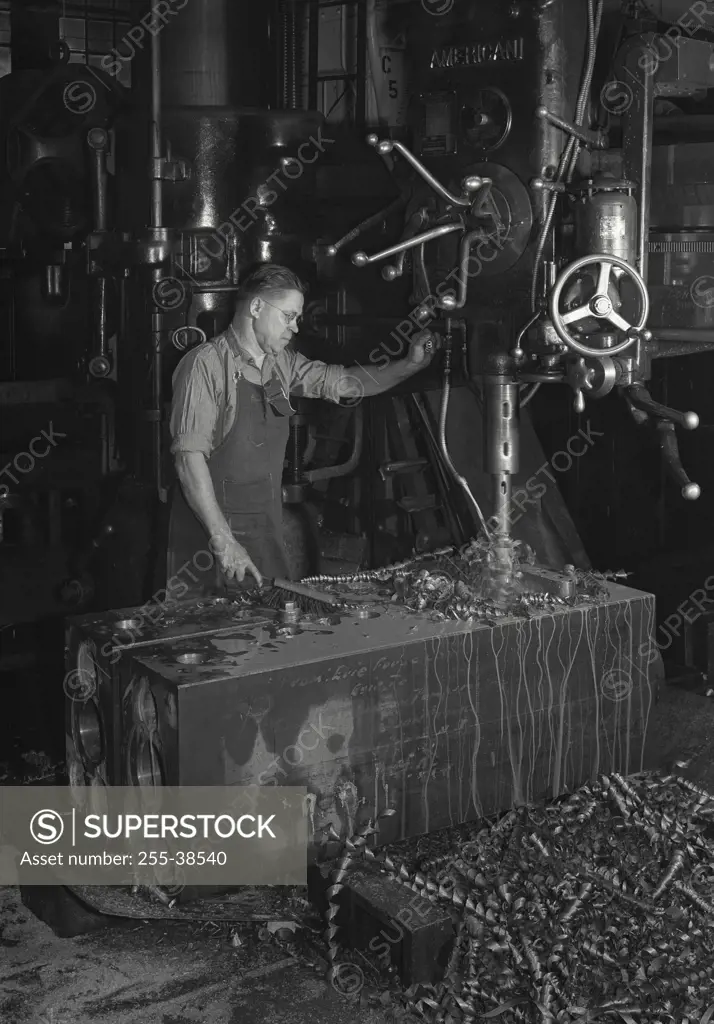 Vintage Photograph. Factory operator drilling through steel block on a Radial Drill Press with soluble oil is running on drill to cool it