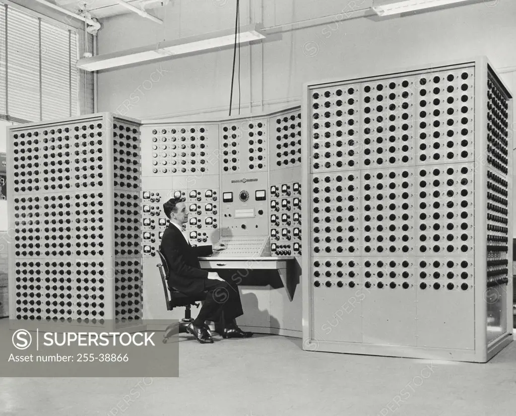Vintage photograph. Man sitting in front of dial studded new computer that mathematically models a complex electric power system.