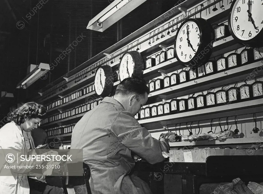 Vintage photograph. Mid adult man and a mid adult woman in a clock factory with clocks on test racks for inspection