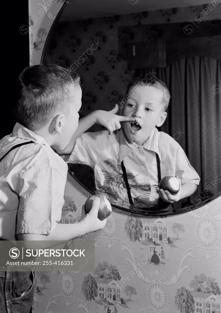 Over shoulder view of boy holding apple, examining gap between teeth in mirror