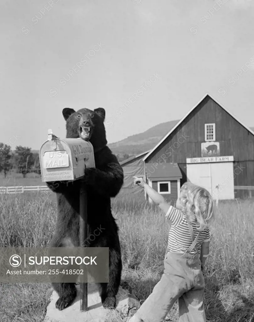 Boy pointing on stuffed bear holding mail box in front of farm building