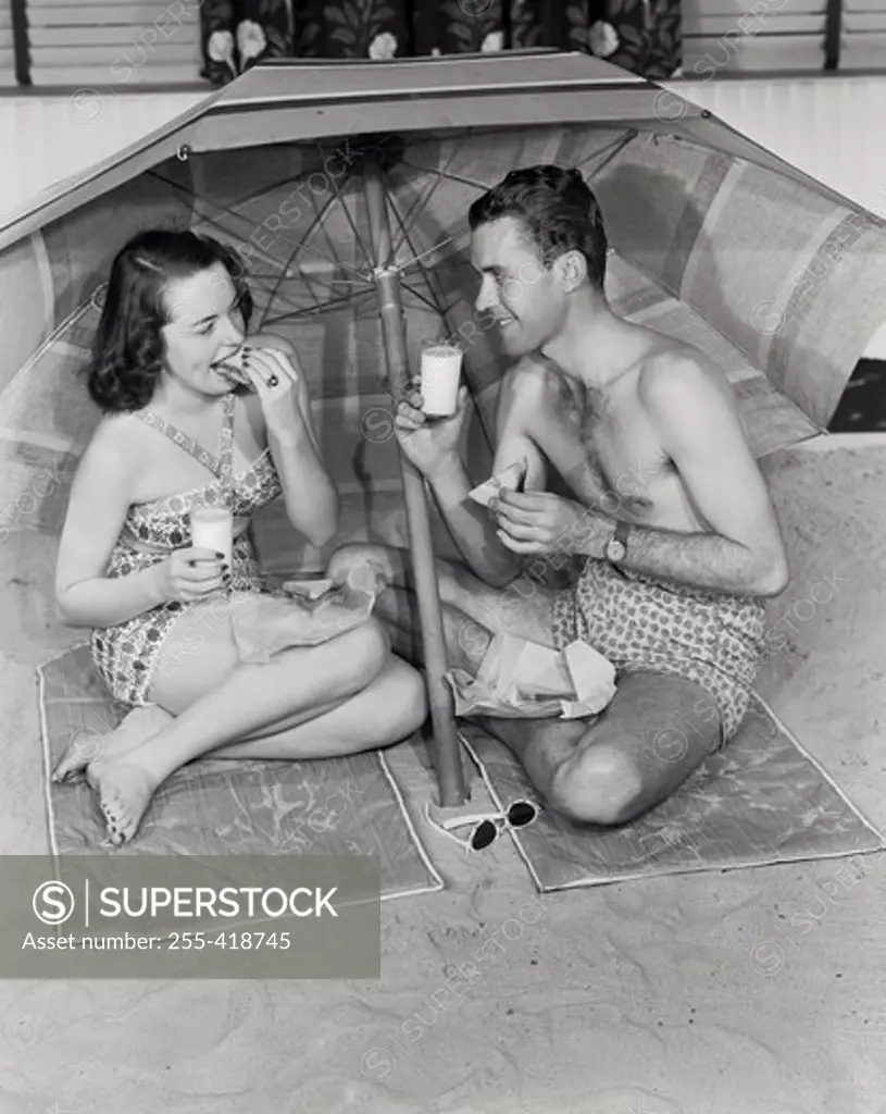 Young couple sitting under beach umbrella, eating snacks