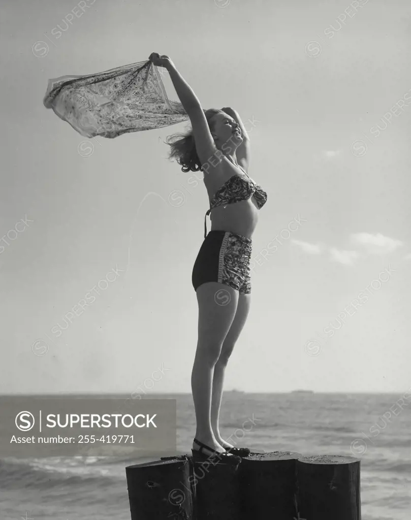 Vintage Photograph. Woman in bikini standing by sea with arms raised and holding scarf