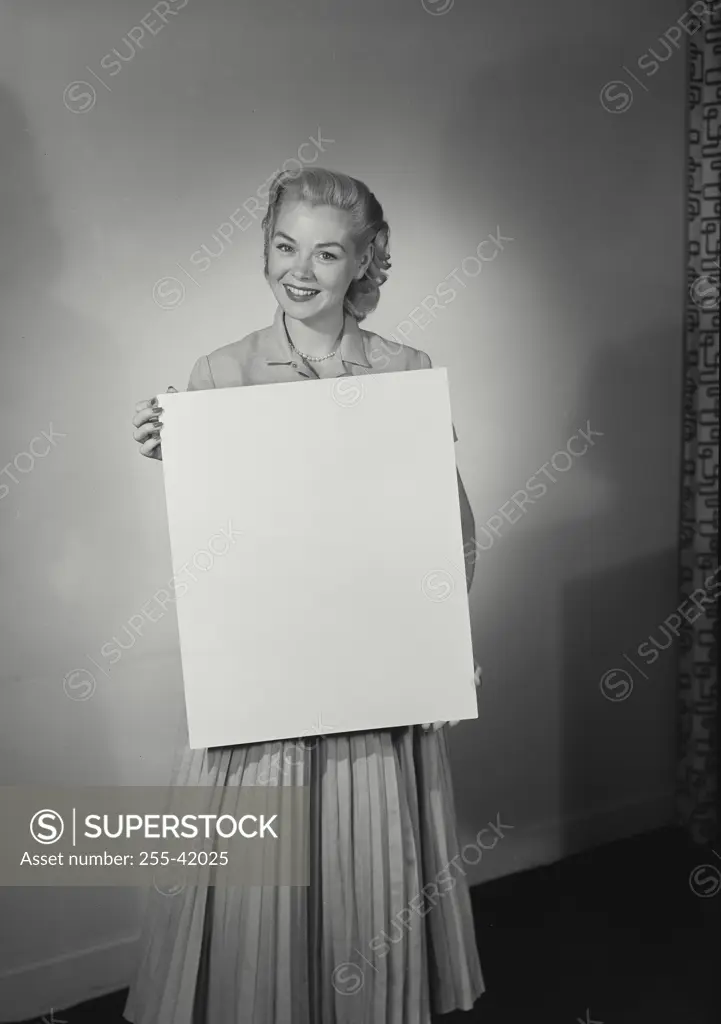 Vintage Photograph. Smiling young woman wearing dress holding up blank white sign in front of wall