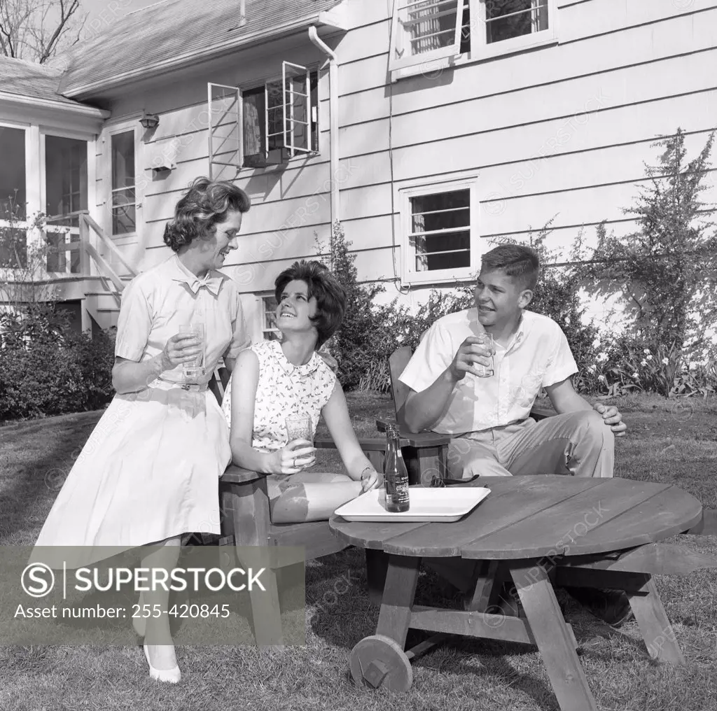 Mature woman with adult son and daughter sitting in backyard and drinking water