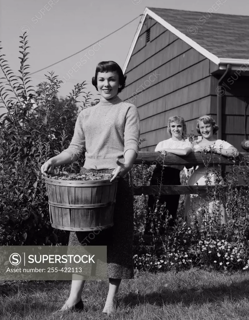 Woman carrying bucket full of dead leaves, two woman watching her and smiling