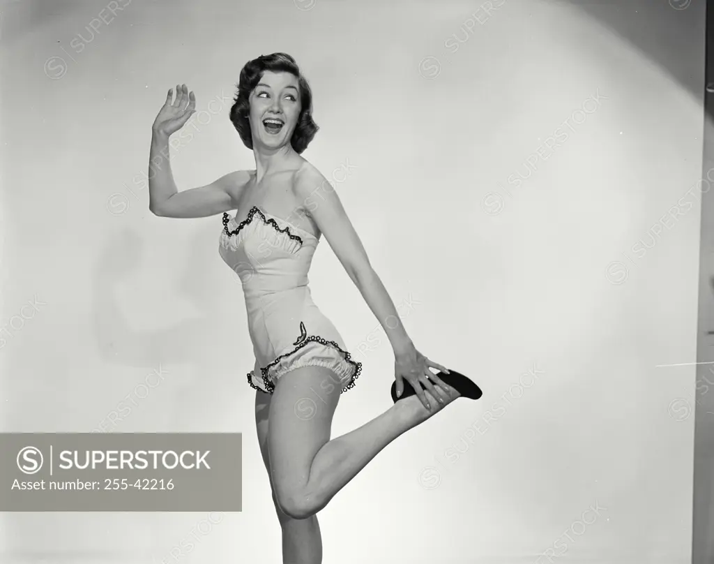 Vintage Photograph. Woman in bathing suit smiling with one arm raised and the other arm touching heel of shoe