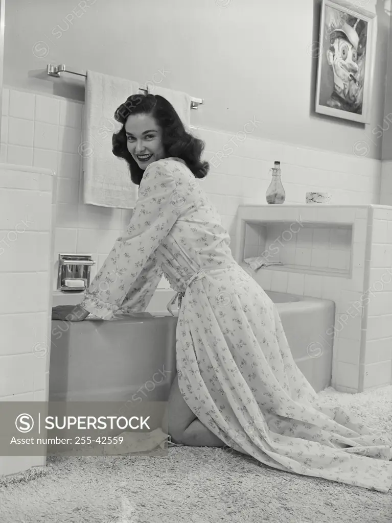 Vintage Photograph. Smiling brunette woman wearing robe kneeling by bathtub in bathroom