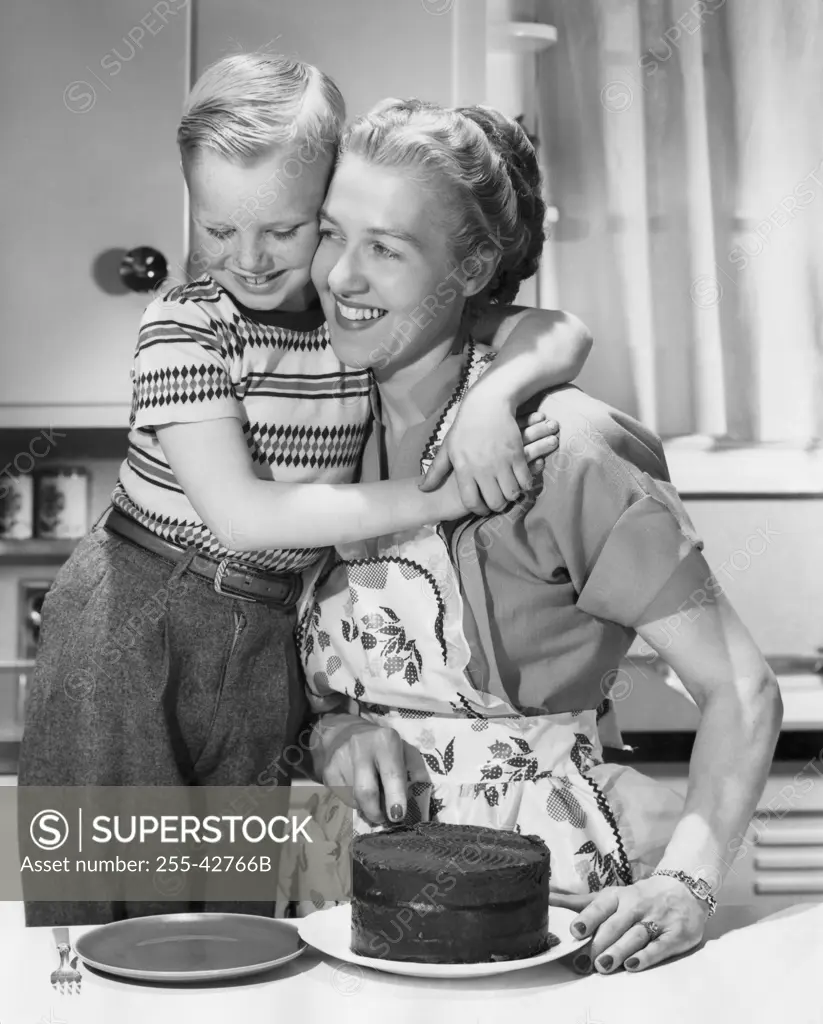 Boy hugging his mother and smiling in front of cake on table