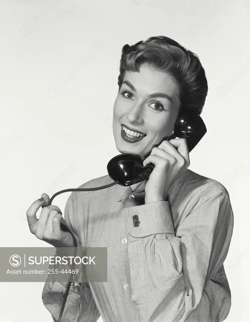 Vintage Photograph. Woman with short hairstyle wearing button up shirt smiling at camera holding up telephone receiver to ear, in conversation