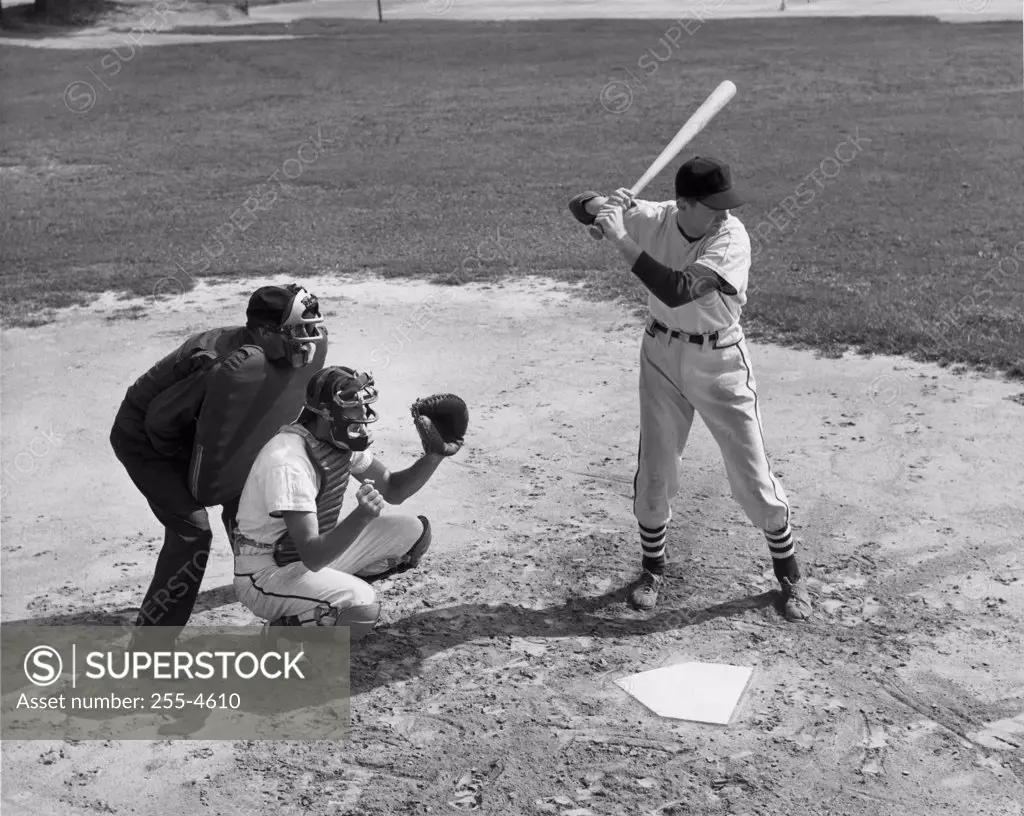 Baseball player swinging baseball bat with baseball catcher and baseball umpire squatting beside him