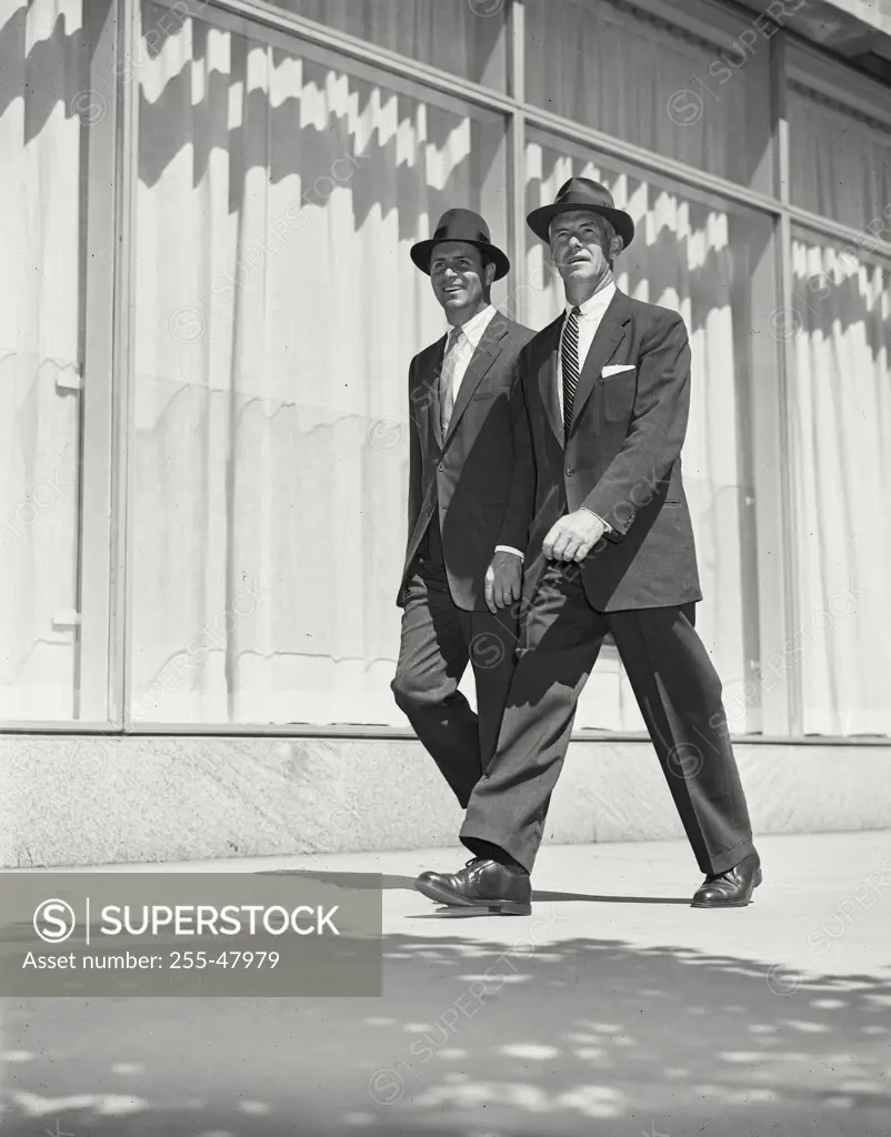 Vintage Photograph. Two men wearing suits and hats walking outside on sidewalk by building with large windows Frame 1