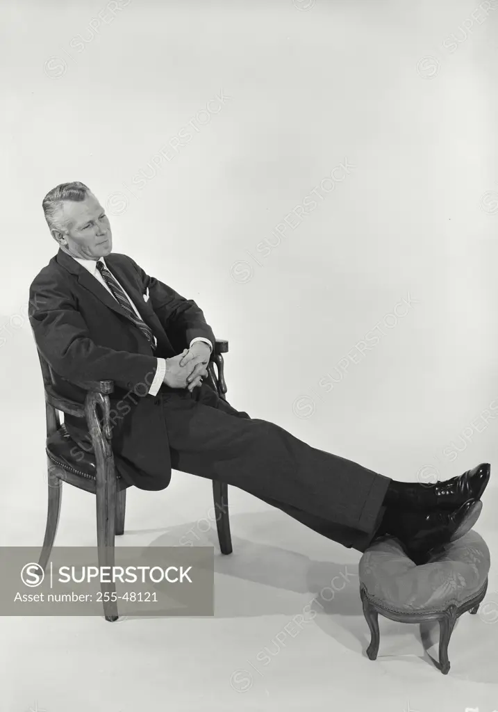 Vintage Photograph. Man wearing suit sitting in chair with feet perched on footstool on white background, Frame 1