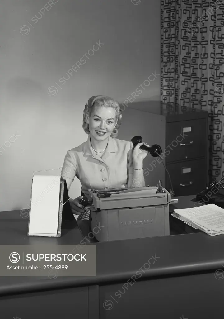 Vintage Photograph. Young woman sitting at office desk with typewriter and holding telephone receiver