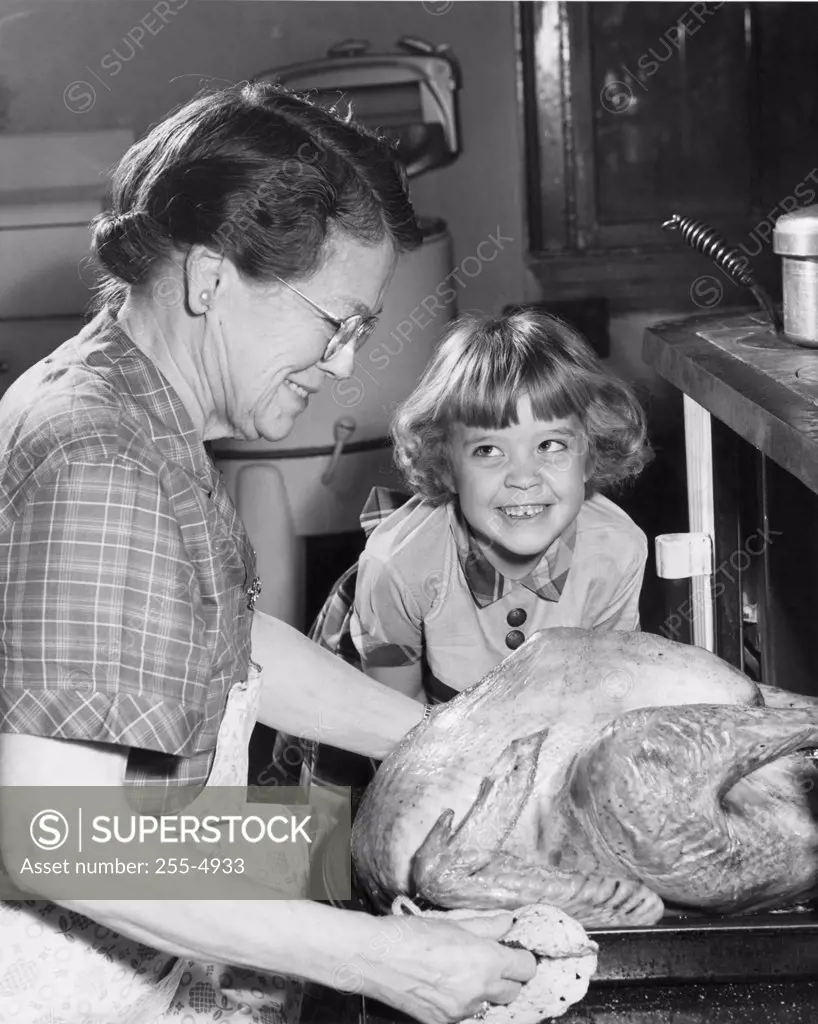 Side profile of senior woman putting roasted turkey in oven with her granddaughter smiling