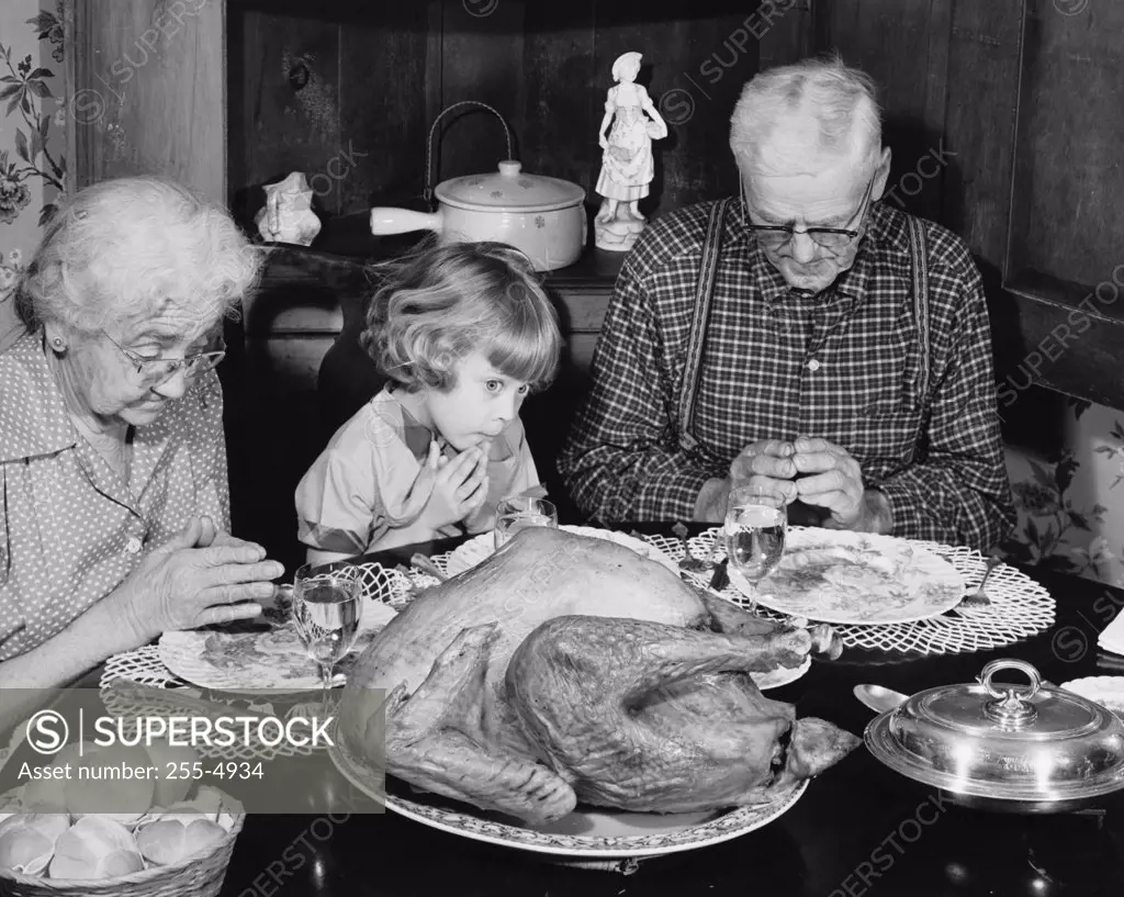 Senior couple praying with their granddaughter at a dining table on Thanksgiving Day