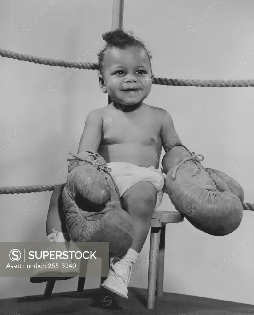 Baby wearing boxing gloves and sitting on stool in boxing ring