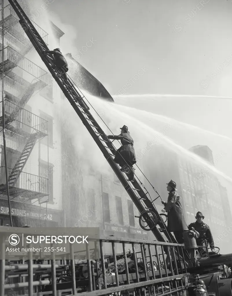 Vintage photograph. Side profile of two firemen climbing a ladder towards a burning building