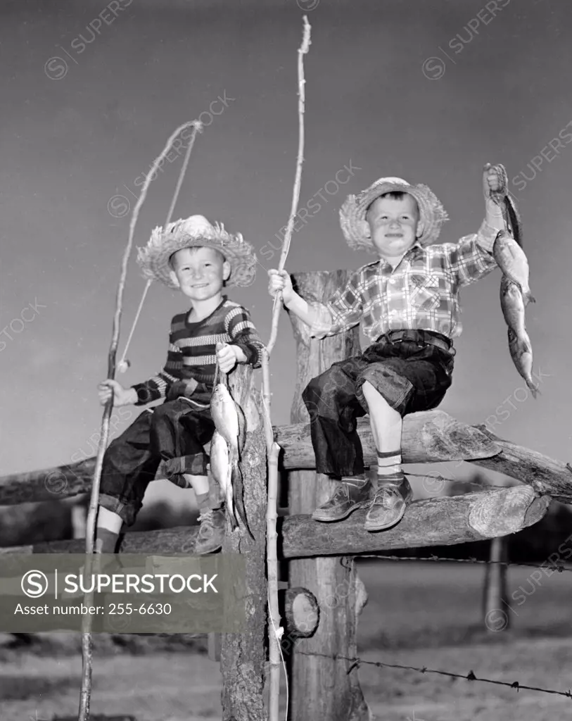 Portrait of two boys with bunch of fish and fishing rods made of sticks sitting on wooden fence