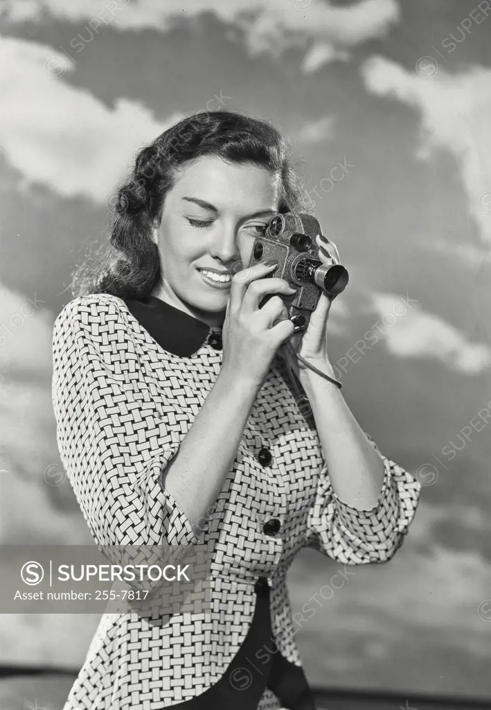 Vintage photograph. Portrait of young woman holding up 8mm movie camera to eye in front of cloud background
