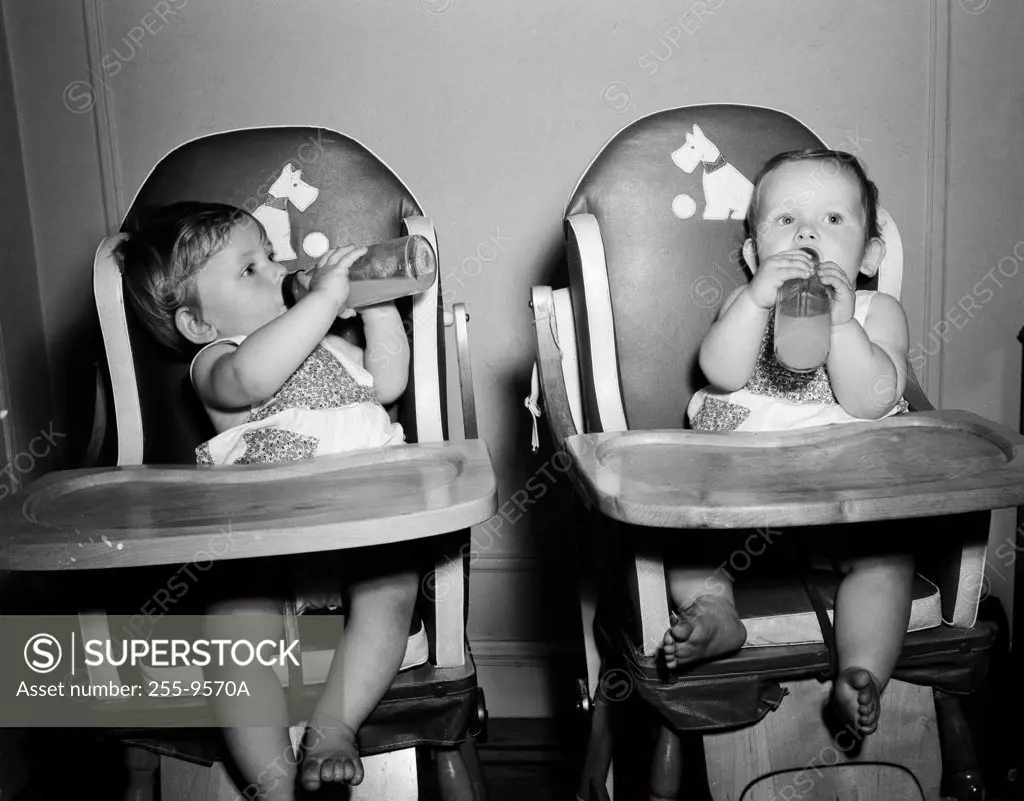 Two babies sitting in high chairs drinking from baby bottles