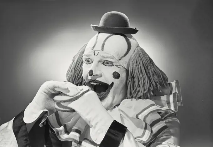 Vintage photograph. Portrait of clown wearing silly hat with hand sandwiched between two pieces of bread.