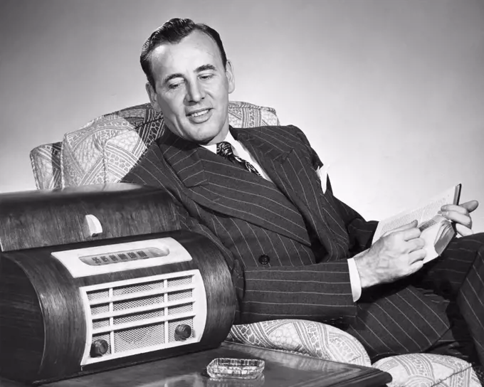 Vintage Photograph. Caucasian man wearing pin striped suit sitting in arm chair looking over at large radio on side table