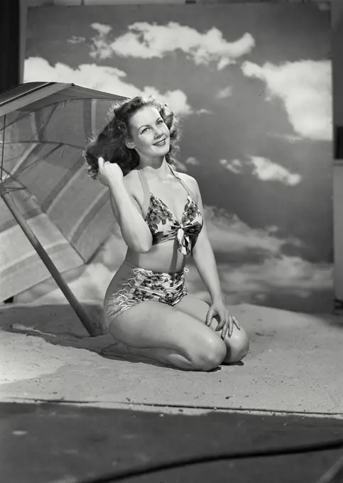 Vintage Photograph. Smiling curly haired woman wearing swimsuit sitting in sand on beach set with large umbrella and sky backdrop playing with hair