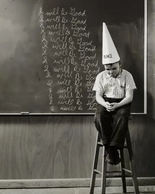 Boy wearing a dunce cap sitting in front of a blackboard