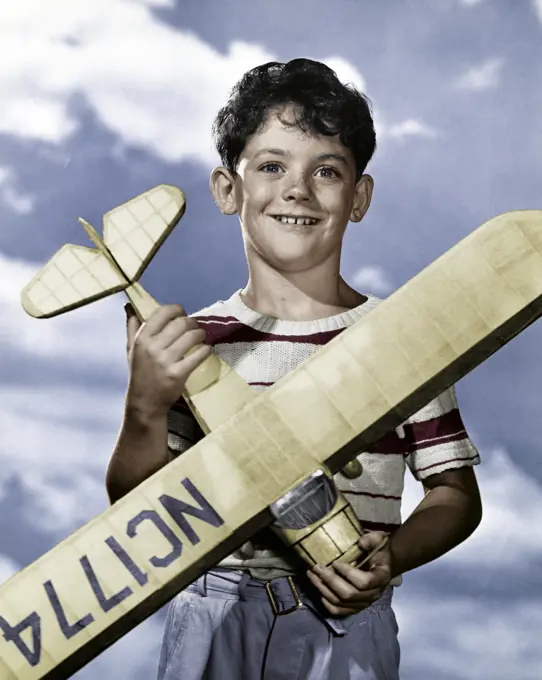 Vintage Photograph. Smiling boy with curly brown hair wearing striped shirt holding large toy airplane in front of cloud sky background