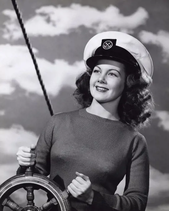 Vintage Photograph. Smiling brunette woman wearing sweater and captain's hat in front of cloud sky background standing with boat helm steering wheel
