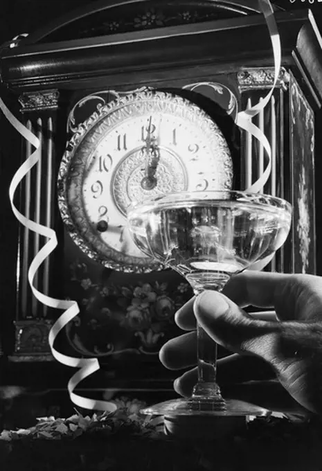 Hand holding champagne flute with champagne, clock showing 12 o'clock in the background