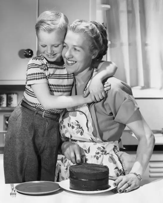 Boy hugging his mother and smiling in front of cake on table