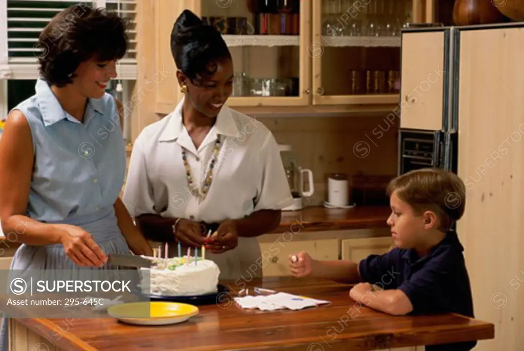Mother cutting a cake with her son and a mid adult woman in a kitchen