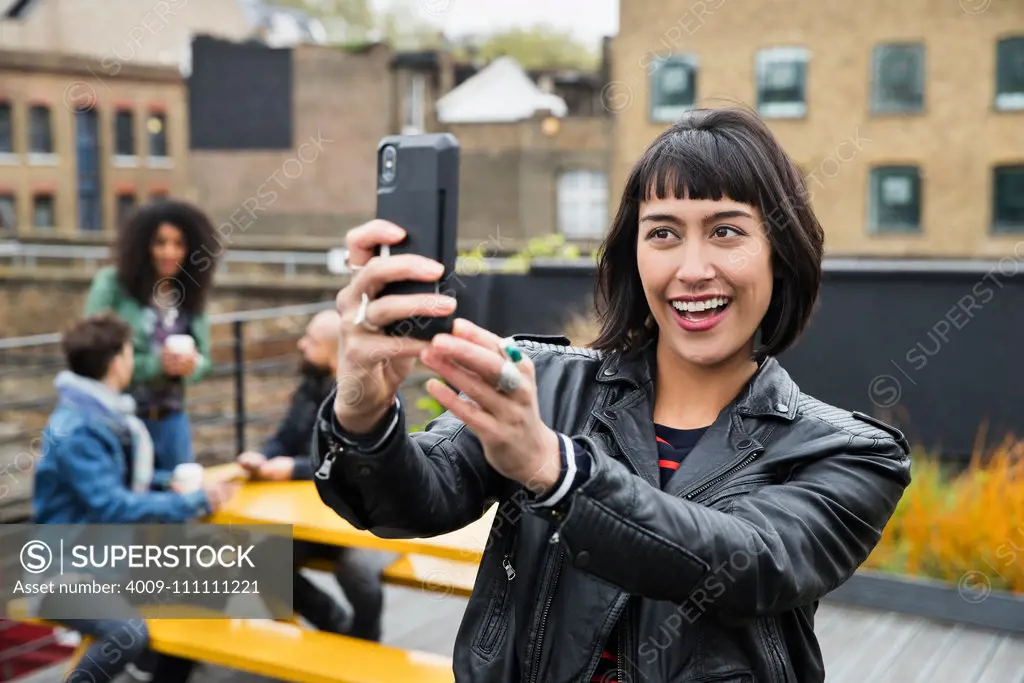 Portrait of Woman taking selfie with group of friends working on tablets at outdoor patio table in co-working space in the background