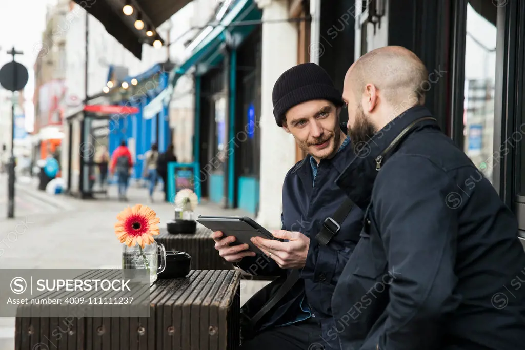 Two men sitting at outdoor cafe table looking at tablet