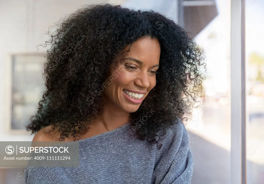 Mixed race, middle-aged woman with natural hair looking out window with a large smile.