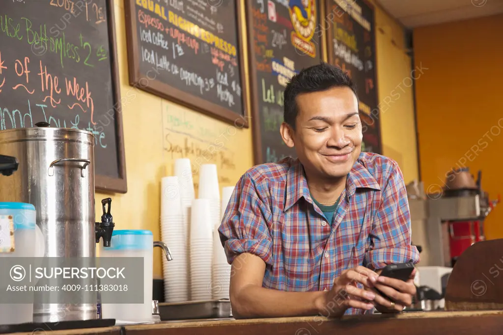 Malaysian business owner standing behind cafe counter with cell phone