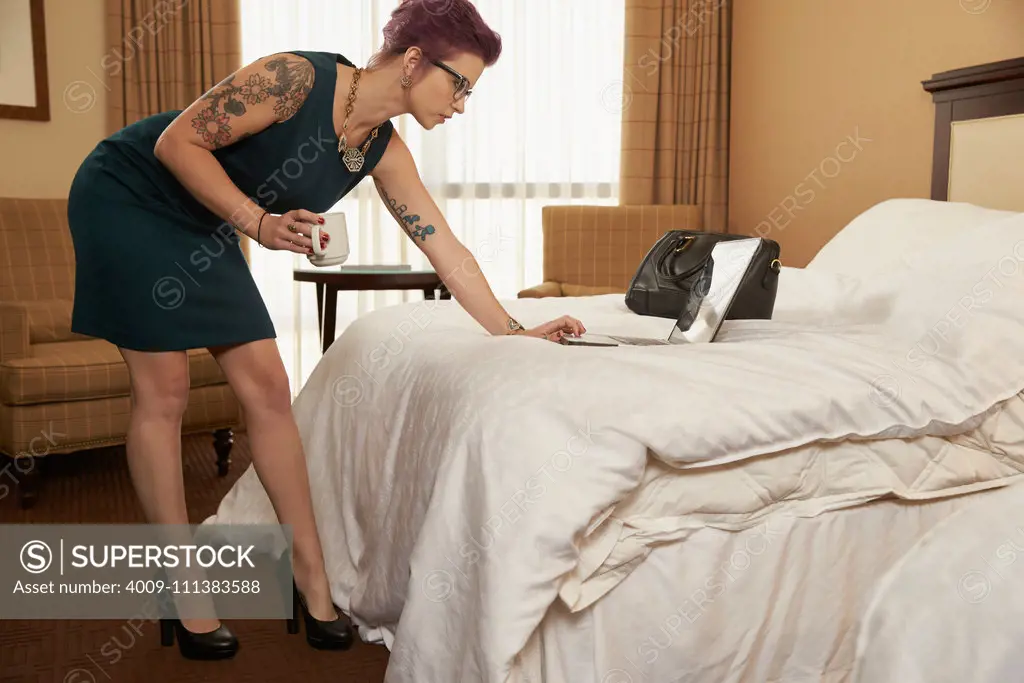 Caucasian woman drinking coffee and using laptop on hotel bed