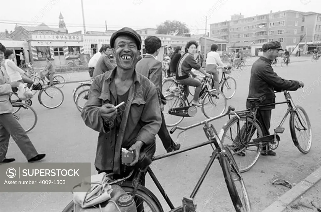 China, Datong, Man with crooked teeth smiling while pulling out cigarette and standing next to his bike, crowd walking and riding behind him