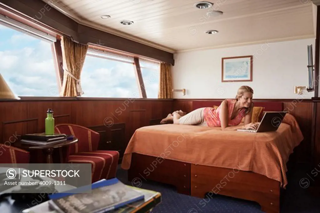 Woman lying on bed using laptop computer in cabin of cruise ship sailing sea