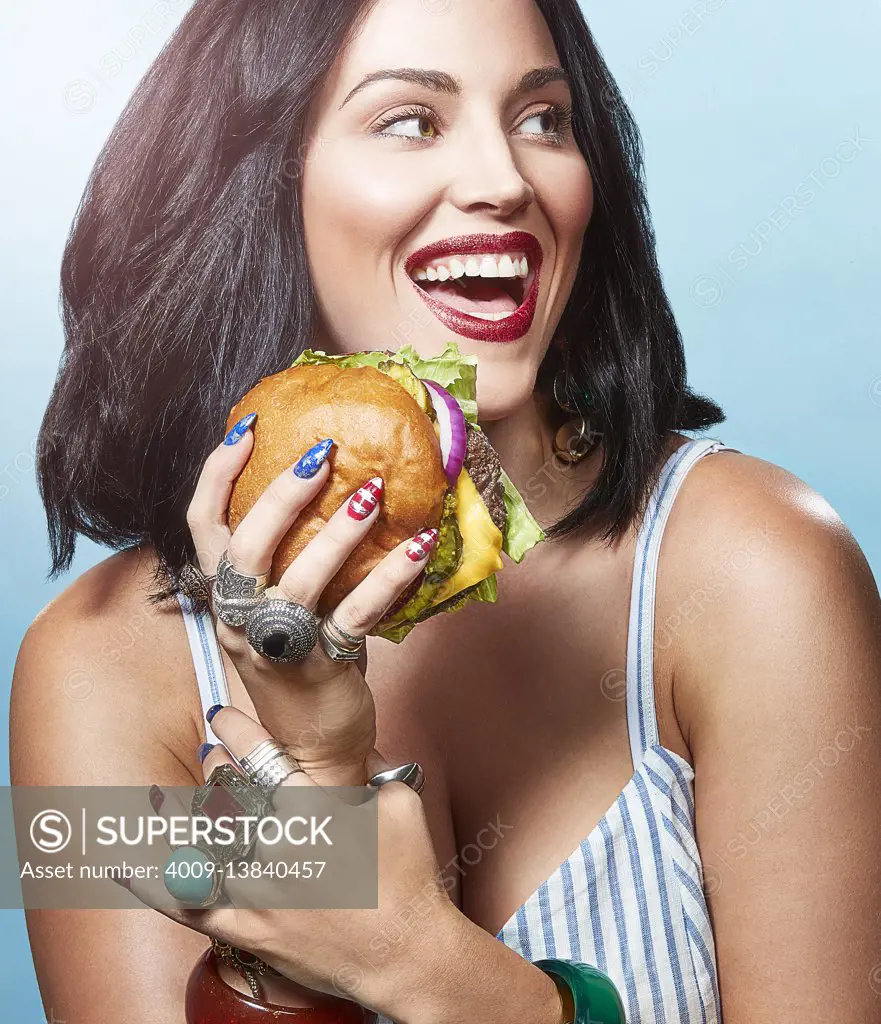A woman in front of a blue background eating a burger