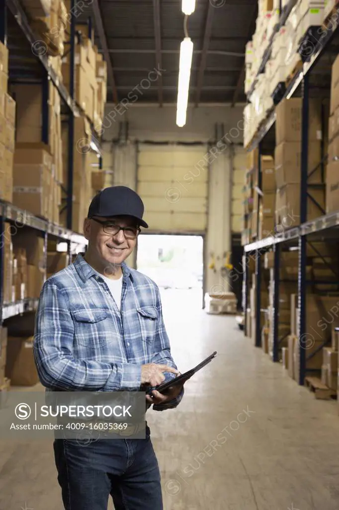 Middle aged warehouse worker smiling at camera and pointing at a tablet
