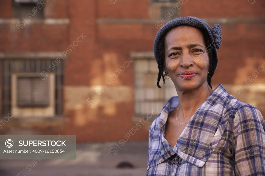 Portrait of older woman wearing knit hat standing in alley smiling looking at camera, brick wall in background  