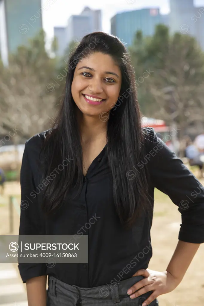 Smiling portrait of young ethnic woman with long black hair standing in park wearing black blouse